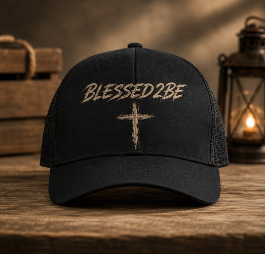 A black trucker-style baseball cap with 'BLESSED 2 BE' and a distressed cross embroidered on the front, displayed on a rustic wooden surface with a warm, atmospheric background featuring stacked wood and a lantern.
