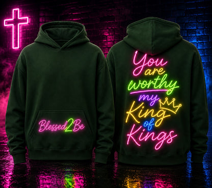 Two dark green hoodies shown front and back against a neon-lit brick wall. The front hoodie has pink and green 'Blessed2Be' text on the pocket area.