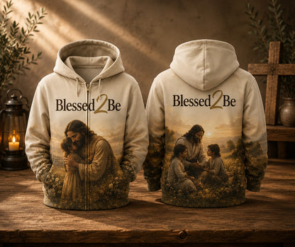 Two hooded zip-up sweatshirts shown front and back on a rustic wooden surface, featuring a warm-toned printed design with the text 'Blessed2Be' and an illustrated scene of Jesus and children. The background includes a lantern, plants, and a wooden cross in a styled indoor setting.