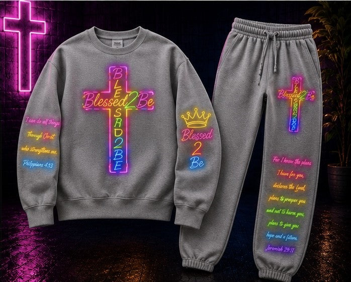 A matching gray sweatshirt and jogger pants set with colorful neon-style cross and crown graphics, plus Bible verse text printed on both pieces, shown against a dark background with a glowing cross sign.