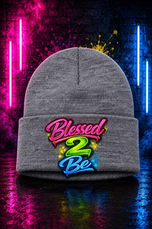 A gray knit beanie with a folded cuff featuring a colorful 'Blessed 2 Be' graphic, set against a dark background with neon pink and blue lights reflecting on a wet surface.