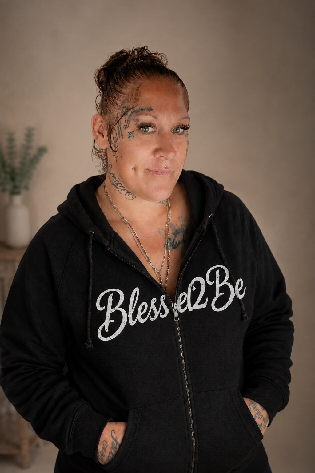 A woman with tattoos wearing a black zip-up hoodie with 'Blessed2Be' printed in white script across the chest, posed against a neutral beige background.